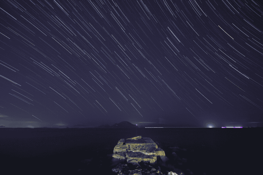 Rastros de estrelas em longa exposição sobre paisagem costeira noturna, técnica relacionada à fotografia de chuvas de meteoros.