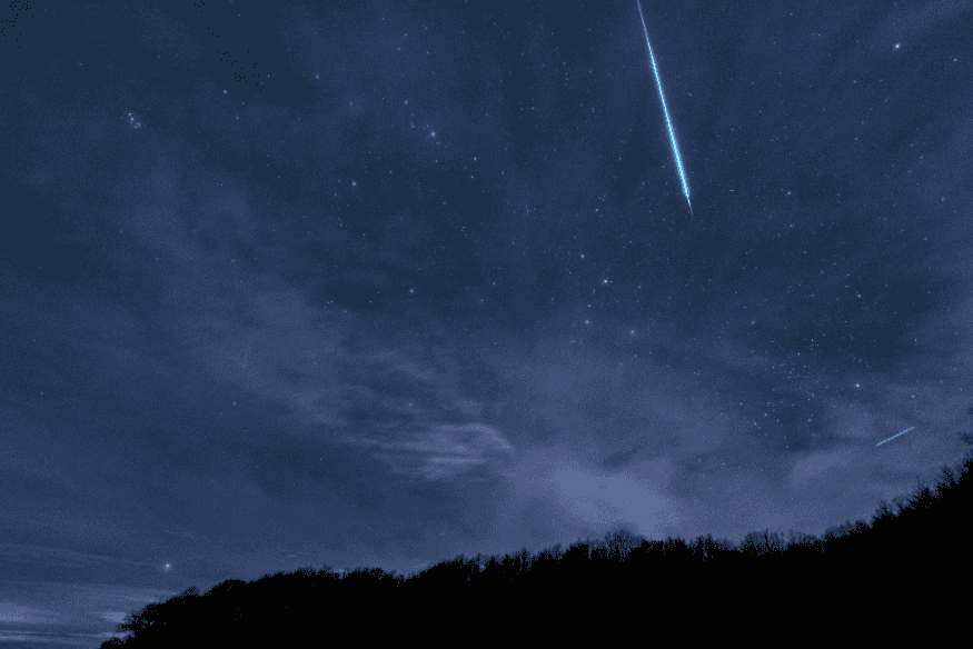 Meteoro brilhante cortando o céu estrelado acima de árvores em silhueta, registrado durante a chuva de meteoros Gemínidas.