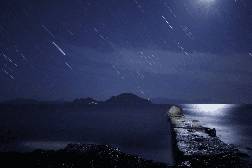 Céu noturno com rastros de estrelas e meteoros sobre o mar, com píer de pedra em primeiro plano, durante as Gemínidas.