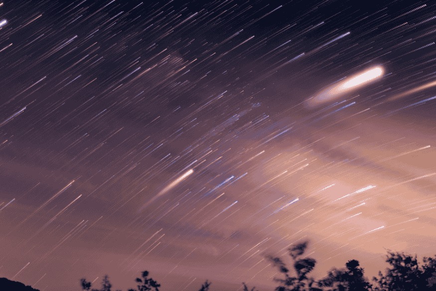 Longa exposição do céu com rastros de estrelas e meteoros brilhantes, com silhueta de árvores no horizonte.