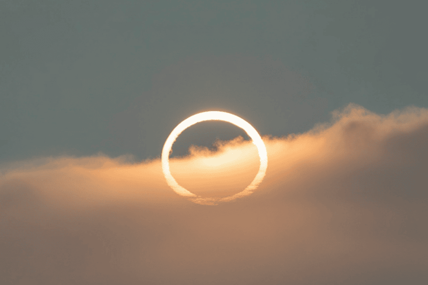 Eclipse solar anular com anel luminoso visível entre nuvens, destacando o contorno circular do Sol.