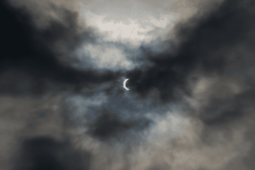 Eclipse parcial pequeno ao centro do céu, cercado por nuvens densas, destacando a mudança de luminosidade durante o evento.