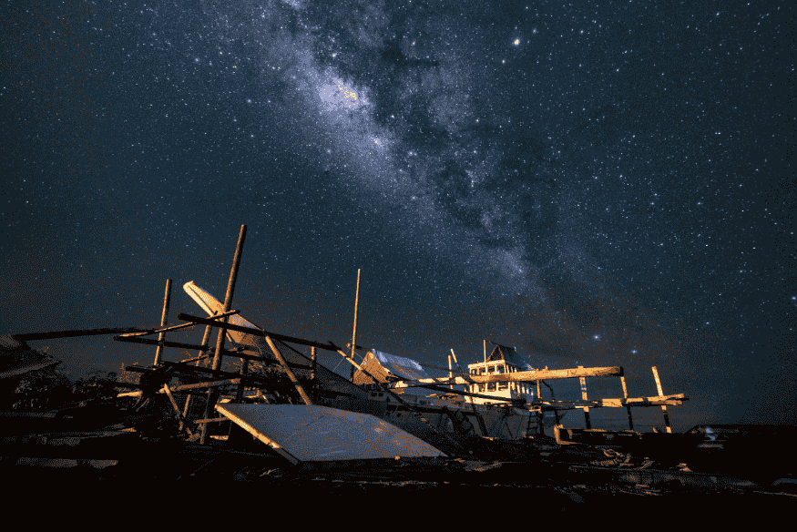 Ruínas de barco antigo sob céu estrelado, simbolizando a relação entre navegação antiga, astronomia e exploração marítima.