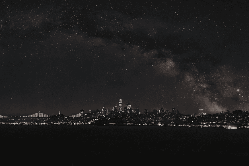 Via Láctea visível sobre o horizonte urbano à noite, ilustrando o contraste entre o céu estrelado e as luzes da cidade.