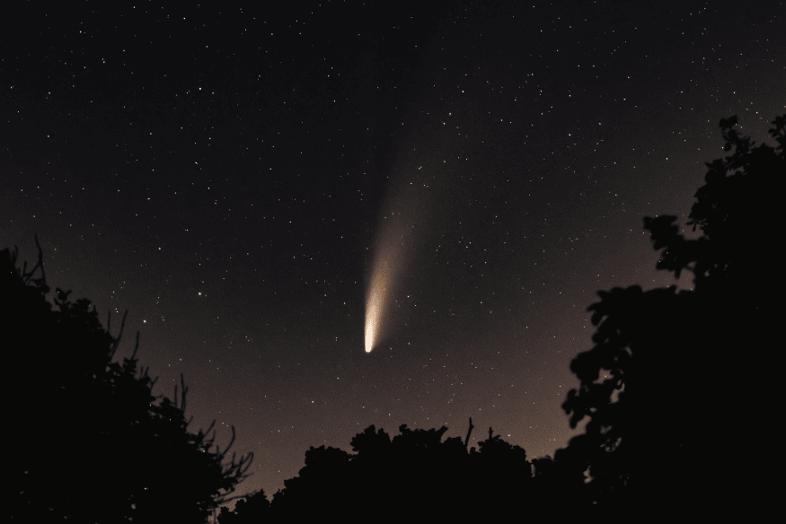Cometa brilhante no céu estrelado acima de árvores, alvo comum para observação e fotografia astronômica com equipamentos adequados.
