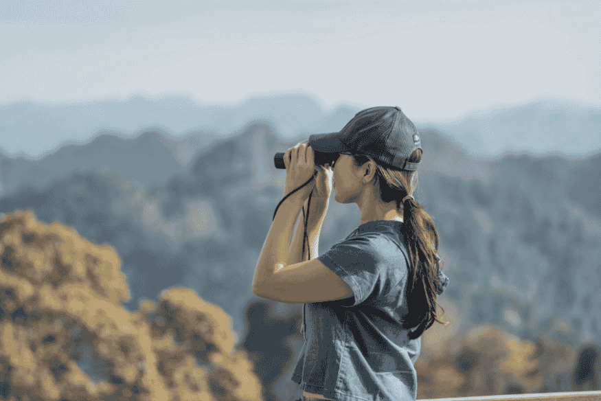 Pessoa usando binóculo em área aberta durante o dia, representando a busca por locais de observação mais adequados e afastados das cidades.