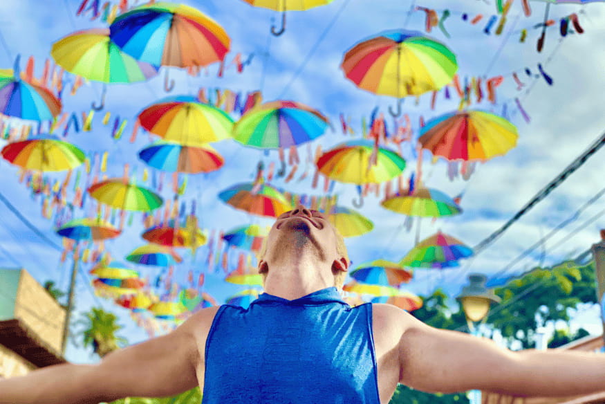 Homem de braços abertos sob decoração de guarda-chuvas coloridos no céu.