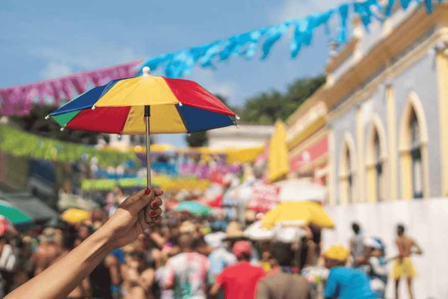Mão segurando sombrinha de frevo com multidão ao fundo nas ladeiras de Olinda.