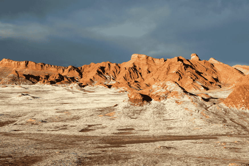Solo esbranquiçado e montanhas alaranjadas no Vale da Morte, em contraste com o céu escuro