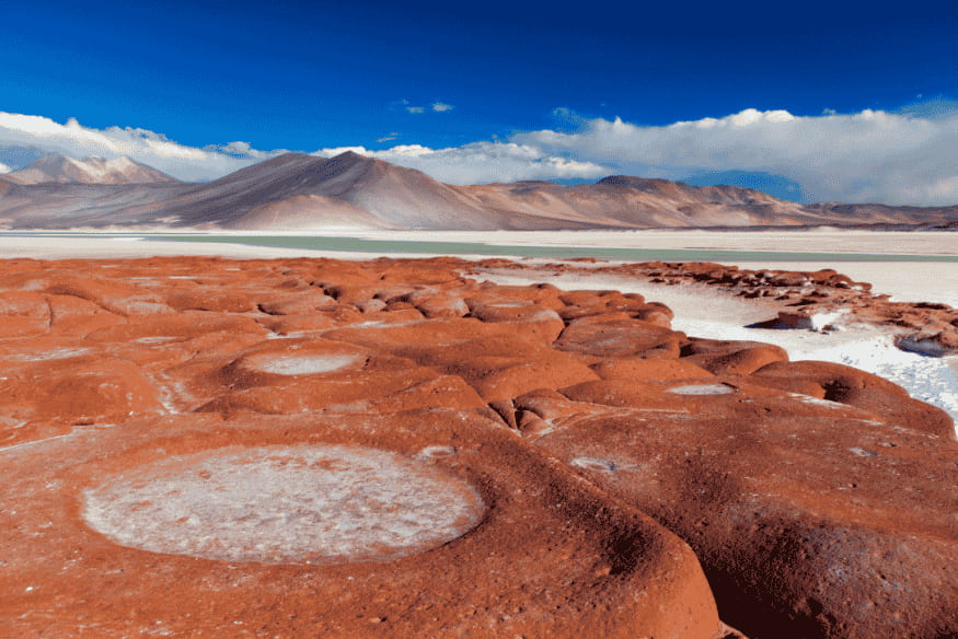 Formações rochosas arredondadas e avermelhadas com manchas de sal, em frente a uma lagoa altiplânica