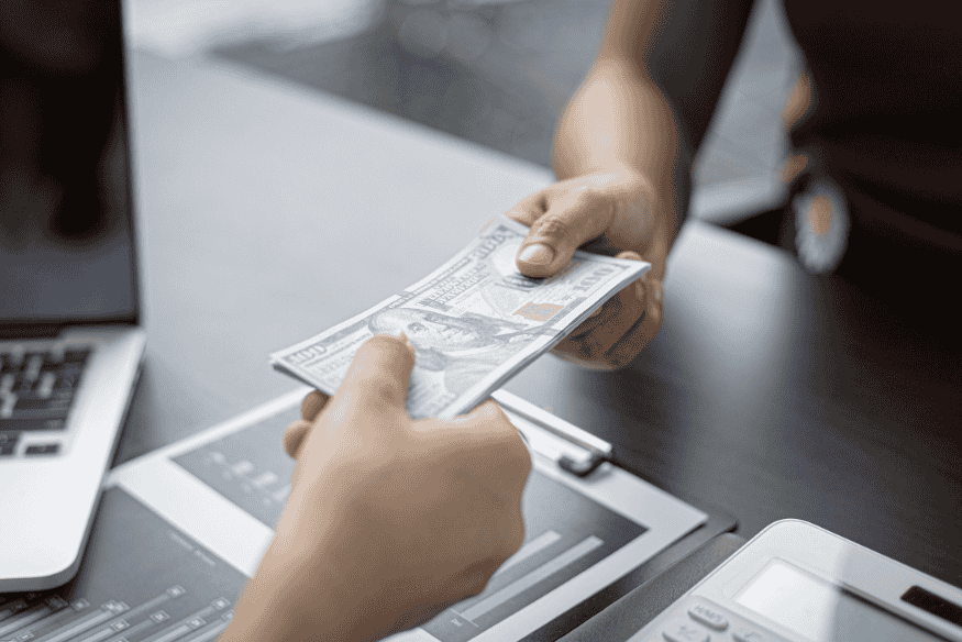 Mão entregando nota de cem dólares a outra pessoa em ambiente comercial