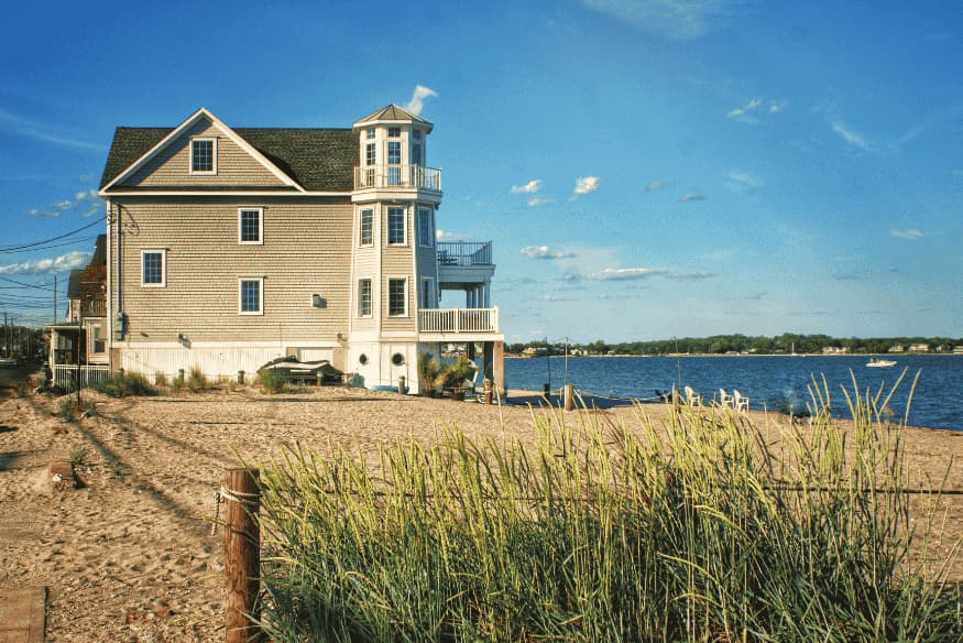 Casa de praia de madeira clara com varanda e vista para o mar em um dia ensolarado.