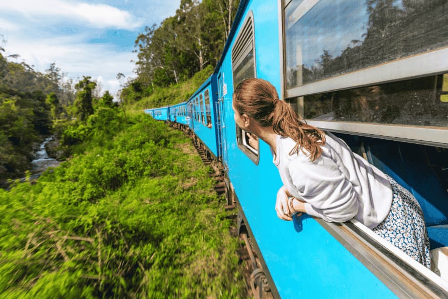 Jovem olhando pela janela de trem azul em trajeto cênico entre montanhas e vegetação