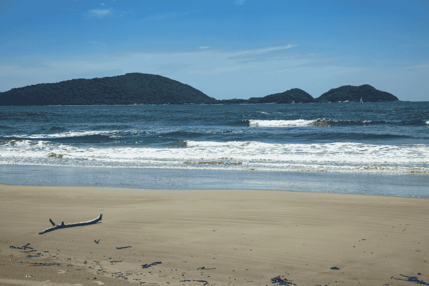 Praia deserta com ondas suaves e galho de madeira na areia, tendo ilhas verdes ao fundo na Ilha do Mel.