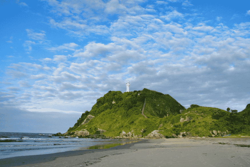 Farol das Conchas visto de baixo, com o mar e a areia em primeiro plano e o céu parcialmente nublado.