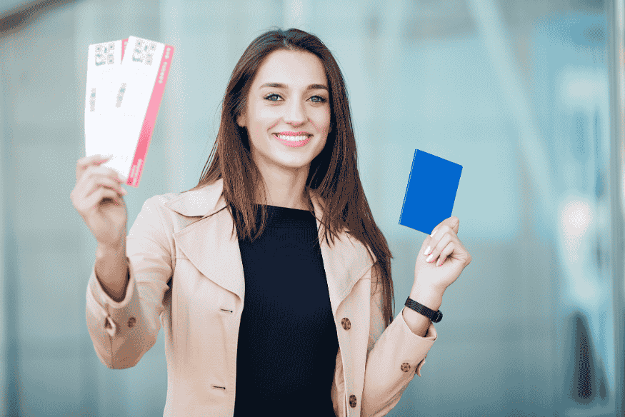 Mulher sorrindo segurando passaporte e passagens aéreas em frente ao aeroporto.