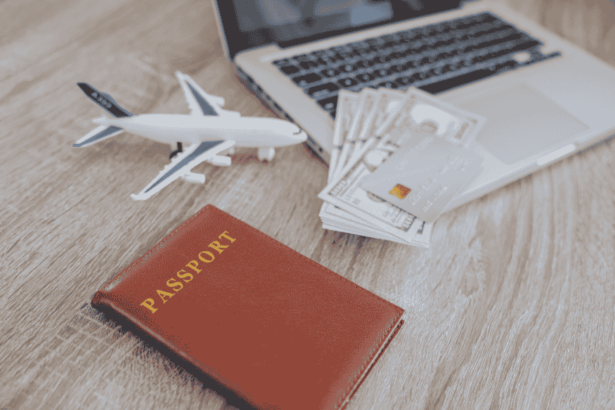 Passaporte, miniatura de avião e notas de dólar sobre uma mesa com notebook.
