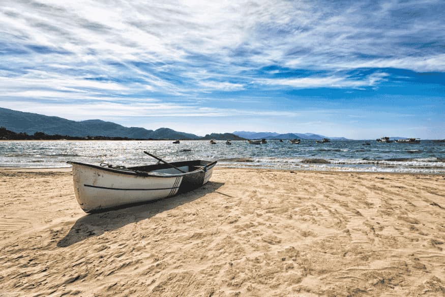 Barco de pesca na areia da Lagoa da Conceição, com montanhas ao fundo e céu parcialmente nublado.