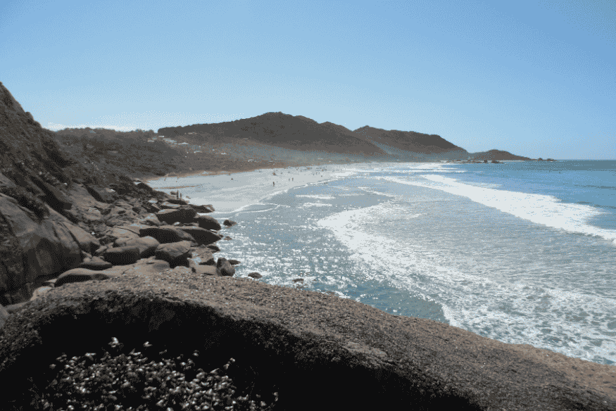 Vista da Praia da Joaquina com ondas fortes e encosta rochosa, procurada por surfistas.