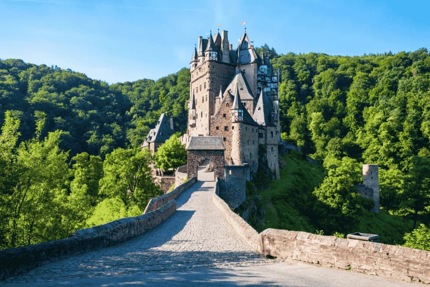 Castelo de Eltz entre colinas verdes e floresta densa, com ponte de pedra ao entardecer.