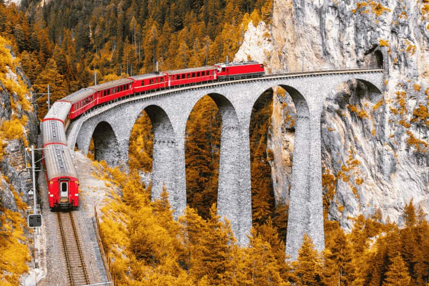 Trem em viaduto curvo sobre penhasco em meio a floresta com folhas amarelas