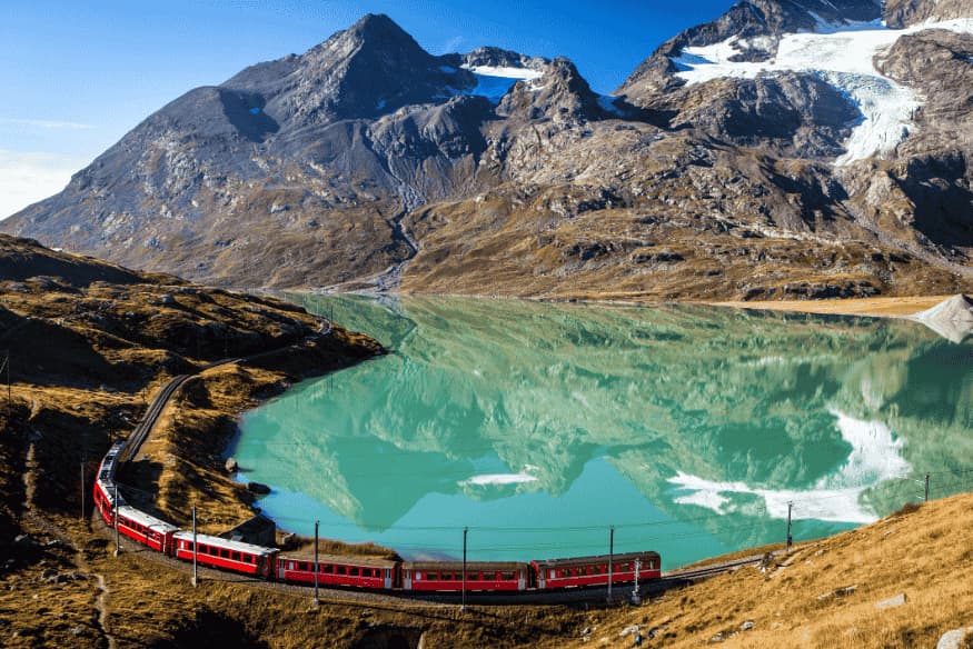 Trem vermelho passando ao lado de lago alpino com reflexo de montanhas nevadas