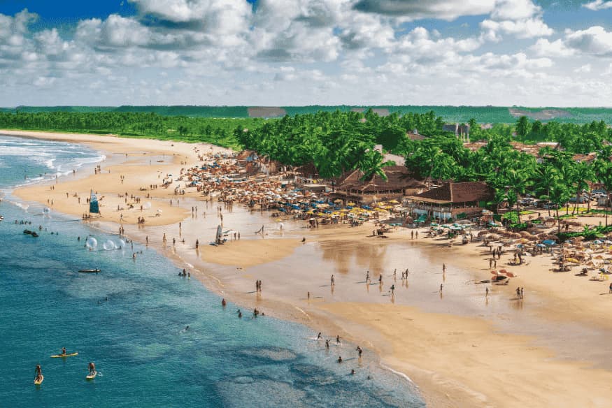 Praia urbana de Maceió com turistas, barracas, mar azul e coqueiros ao fundo