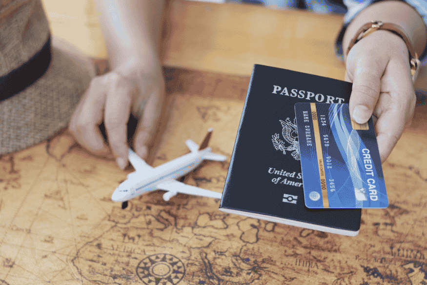 Pessoa segurando passaporte e cartão de crédito ao lado de miniatura de avião sobre mapa-múndi.