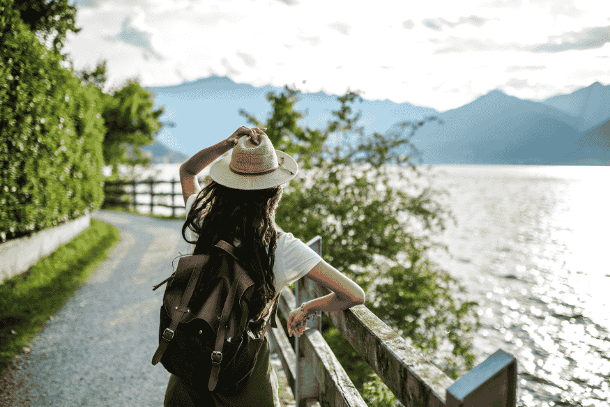 Mulher com mochila e chapéu caminhando sozinha à beira de um lago cercado por montanhas.