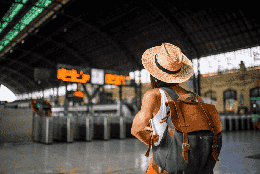 Mulher de chapéu e mochila observando o painel de horários em uma estação ferroviária.