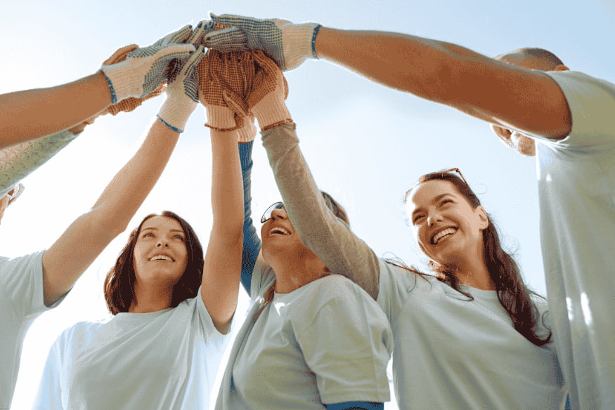 Voluntários de mãos dadas comemorando em equipe sob céu azul.