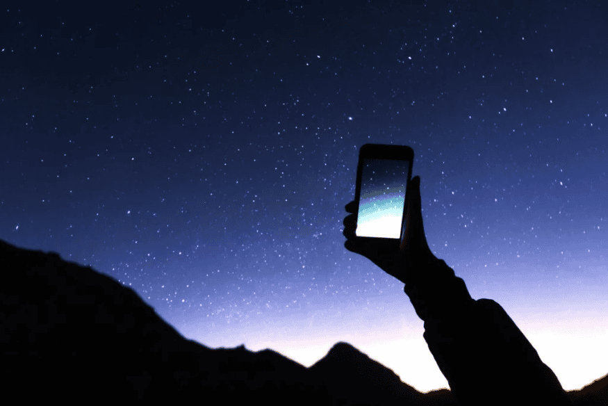 Pessoa segurando celular apontado para o céu estrelado, usando aplicativo de astronomia para localizar estrelas e constelações.
