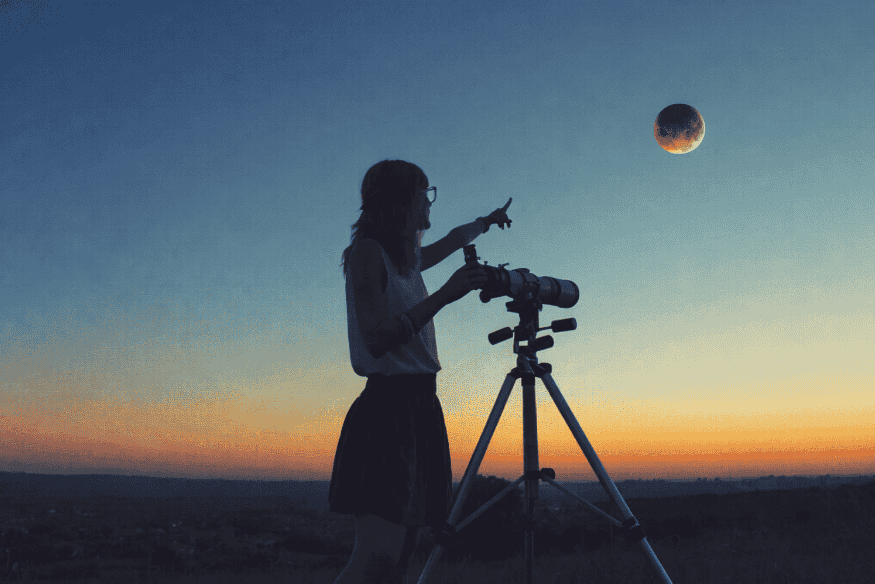 Silhueta de pessoa com câmera em tripé apontando para a Lua eclipsada no céu ao entardecer, em sessão de observação astronômica.