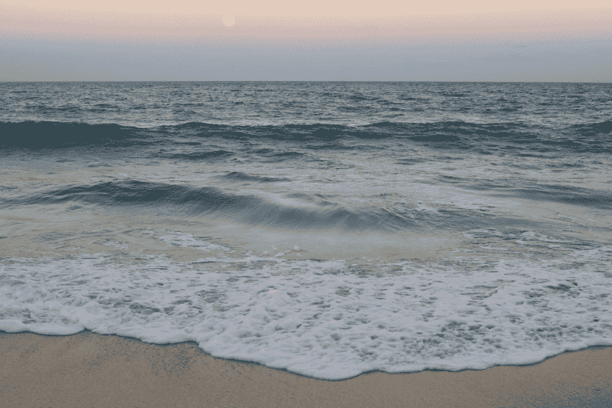 Ondas suaves chegando à praia com espuma na areia e horizonte do oceano, imagem de referência para variação de marés.