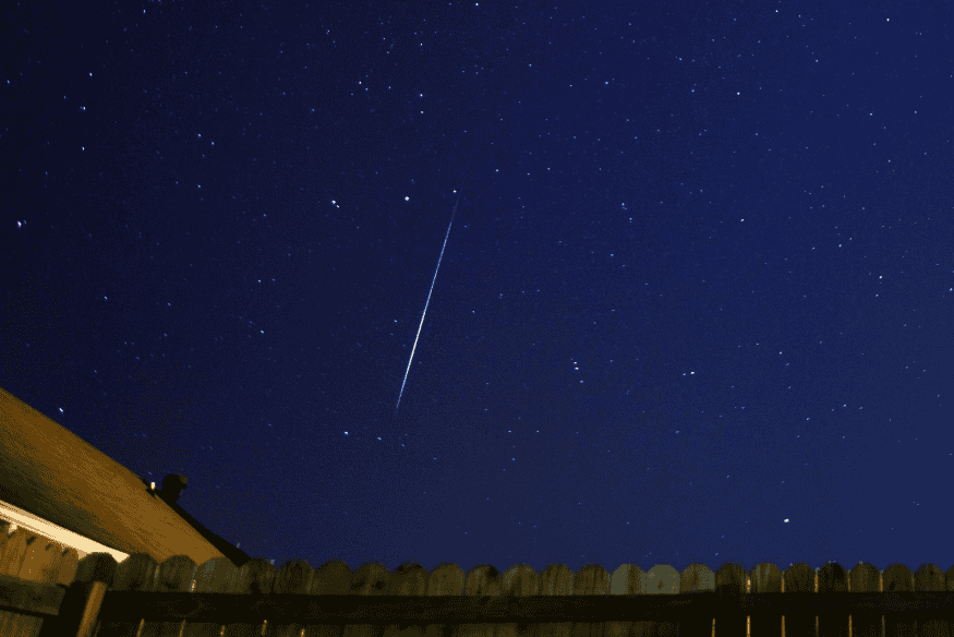 Meteoro riscando o céu estrelado acima de casas e cerca, registro típico de chuva de meteoros.
