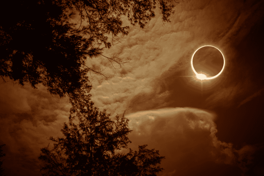 Eclipse solar anular com “anel de fogo” no céu, visto entre nuvens e silhuetas de árvores ao redor.