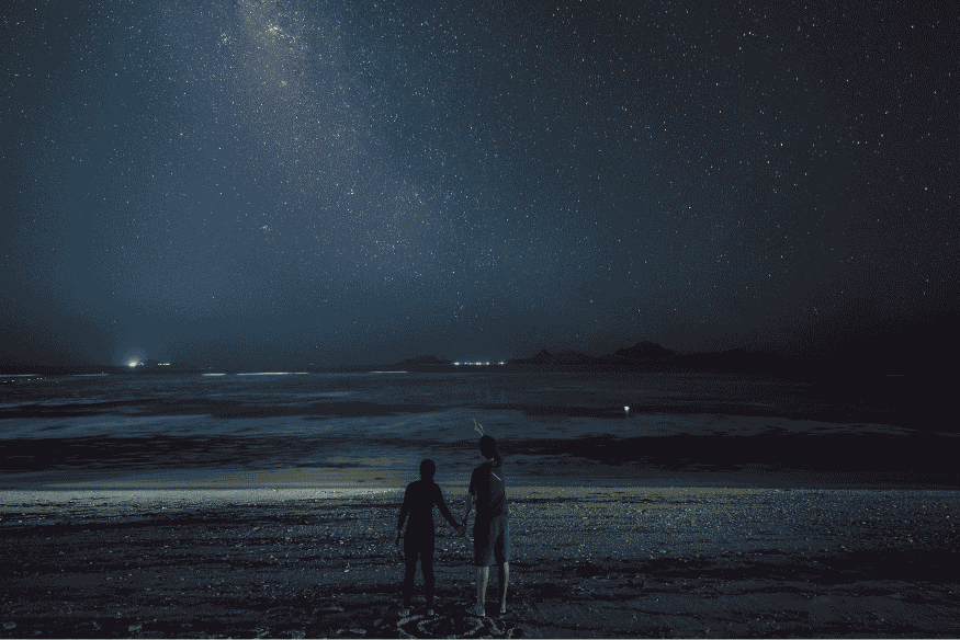 Duas pessoas observando o céu estrelado na praia à noite, cenário ideal para ver planetas visíveis a olho nu.